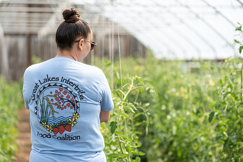 Stephanie Dodge in a greenhouse. The back of their shirt says: Great Lakes Intertribal Food Coalition