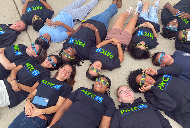 Teenagers wearing sunglasses and PATCH t-shirts lie on the ground with their heads together