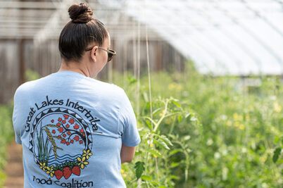 Stephanie Dodge in a greenhouse. The back of their shirt says: Great Lakes Intertribal Food Coalition