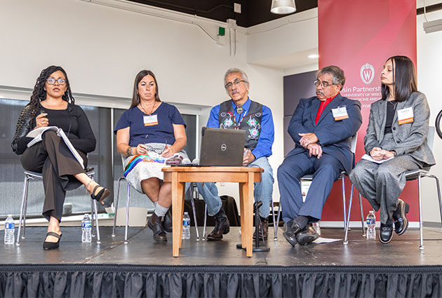 Six panelists sit and listen together while one speaks into a microphone