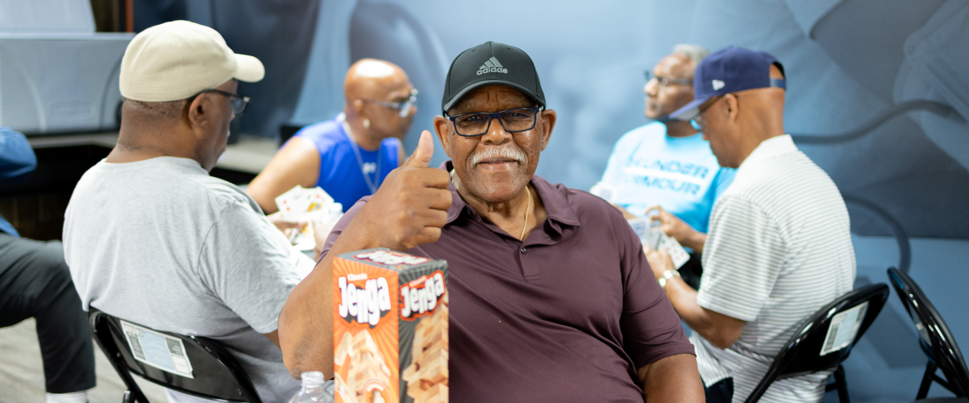 A man shows a thumbs up while others play cards in the background